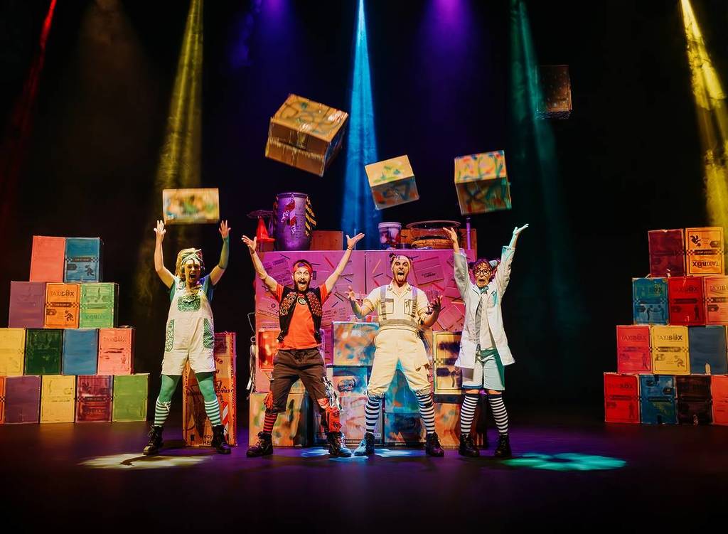 Performers from The Box Show on stage, lit by colourful stage lights.