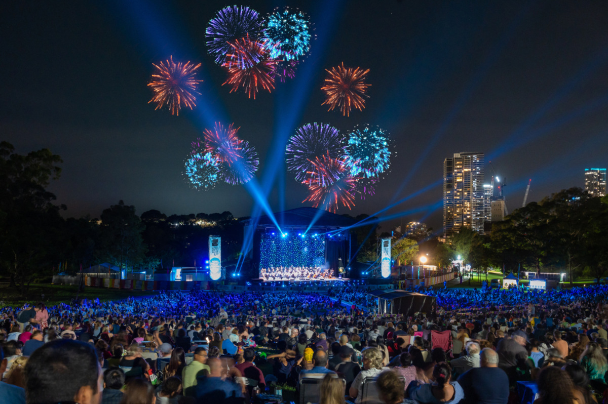 Fireworks illuminate the night sky above a stage as musicians perform during Symphony Under the Stars for sydney festival with a large crowd of spectators