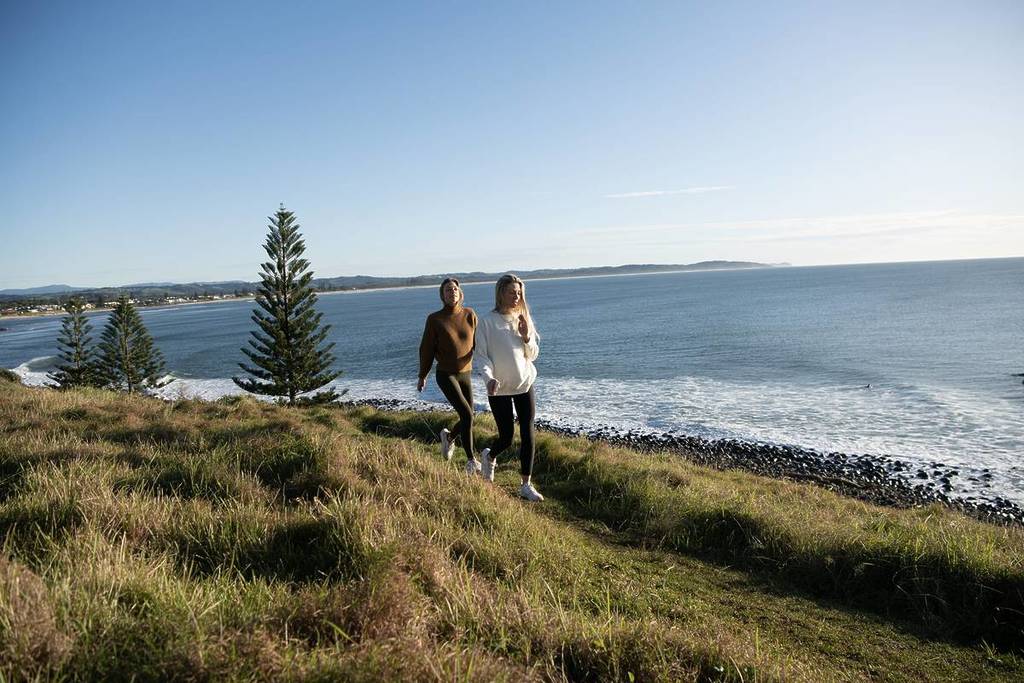Two friends walking up a trail toward Pat Morton Lookout in Lennox Head, surrounded by sweeping coastal views on a crisp winter day
