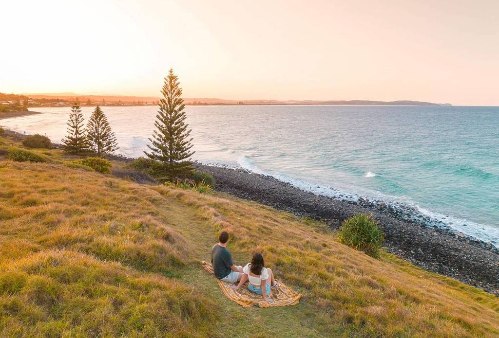 A couple sitting at Pat Morton Lookout in Lennox Head, NSW, with coastal views in front of them and an orange sunset sky overhead