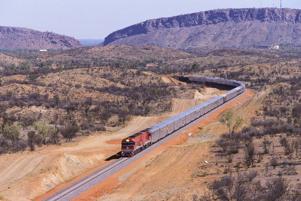 The Ghan train winding through the rugged red landscape of the Australian outback, with rocky hills and sparse bushland in the background