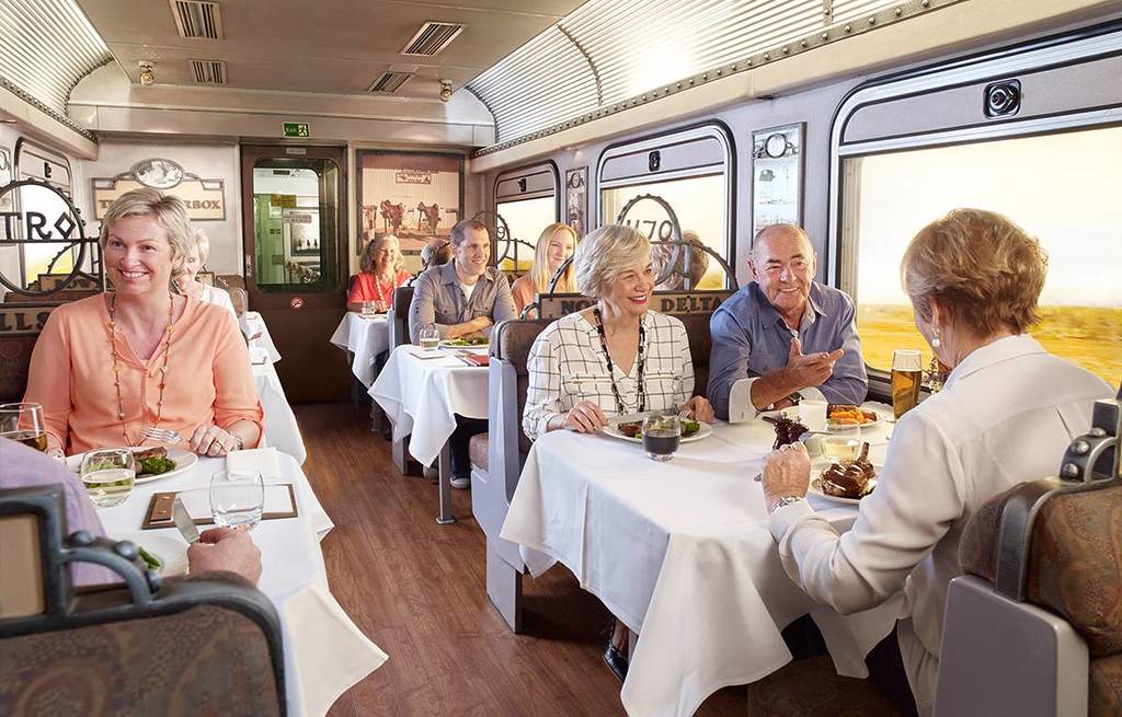 Passengers dining inside a luxury train carriage, enjoying a meal with white tablecloth service, large windows, and scenic views of the Australian landscape outside