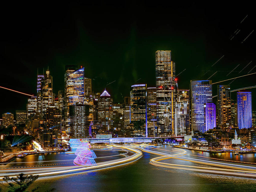 Sydney Harbour at night, with light trails from ferries, illuminated skyscrapers, and the Harbour Bridge glowing in the background
