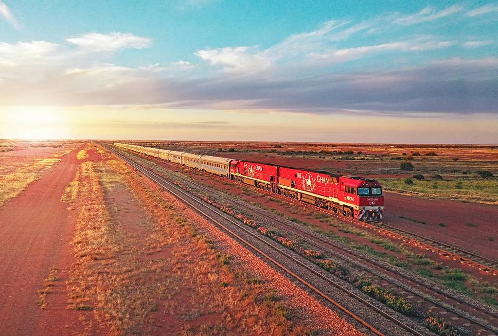 A red Ghan train travels through the Australian outback at sunset, with long carriages stretching into the distance under a wide sky of soft pink and blue hues