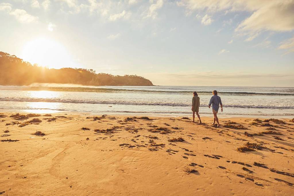 Two people walking along McKenzies Beach at sunset, with golden light reflecting off the sand and water.