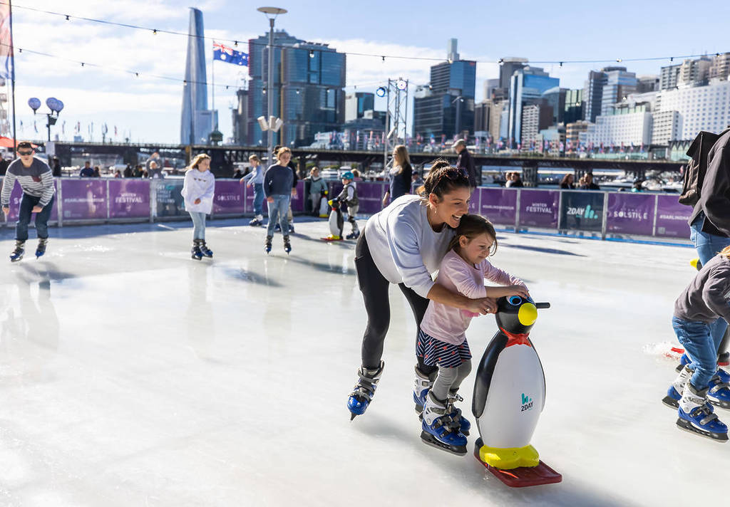 A woman on ice skates pushes along a small child holding a penguin skate aid, at the Darling Harbour winter ice rink.