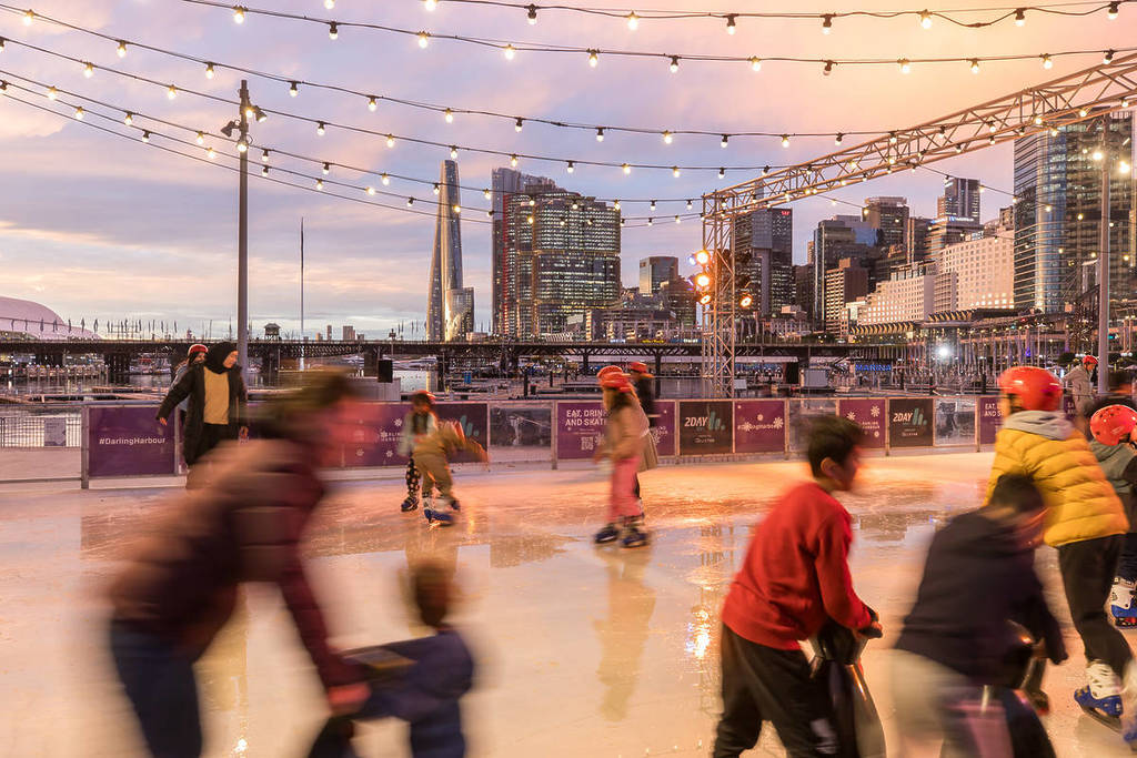 Skaters twirl around the Darling Harbour ice rink at sunset, with the CBD skyline visible in the background.