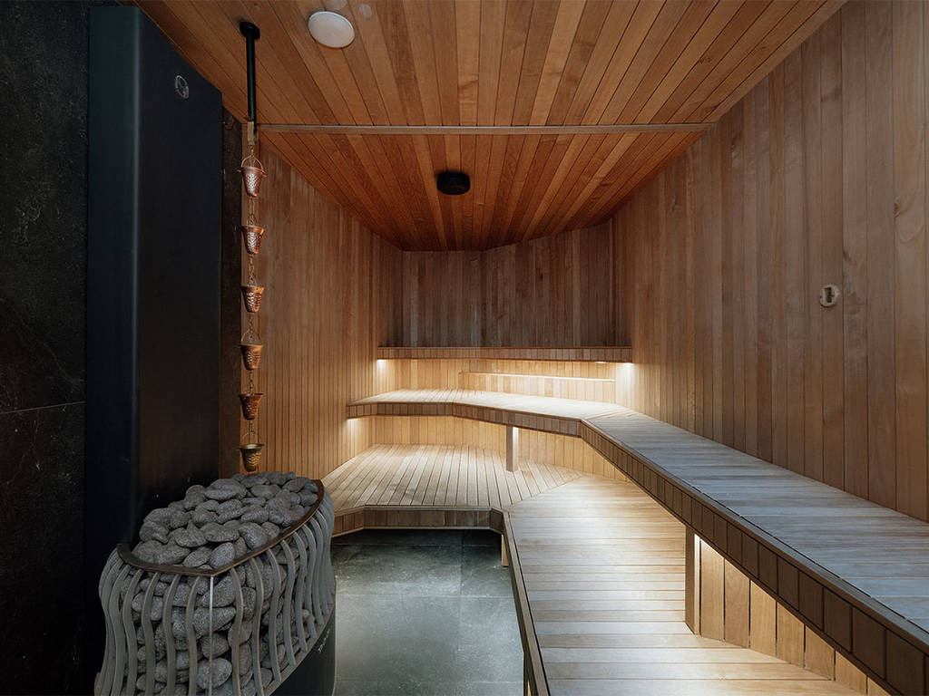 A modern timber-lined sauna with tiered bench seating, soft under-seat lighting, and a stack of smooth sauna stones beside a copper ladle chain at Aqua Ignis bathhouse