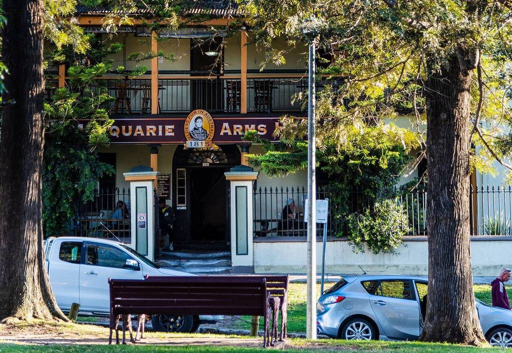 Street view of the Macquarie Arms Hotel in Windsor, framed by tall trees with cars and people outside the historic pub