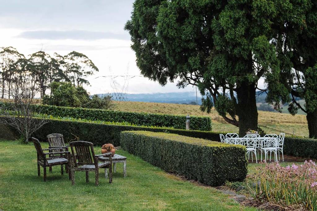 A serene garden with white outdoor chairs and tables set beneath tall trees, bordered by trimmed hedges and open farmland in Robertson, NSW