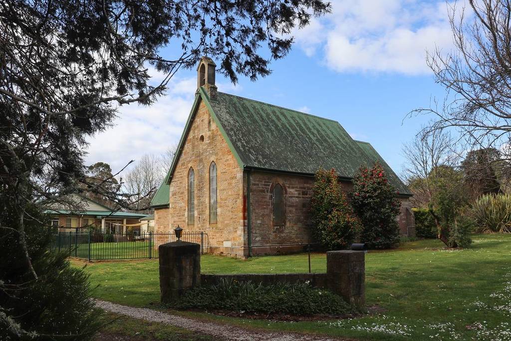 A historic sandstone church with a mossy green roof, surrounded by flowering bushes and a neatly kept lawn in Robertson, NSW