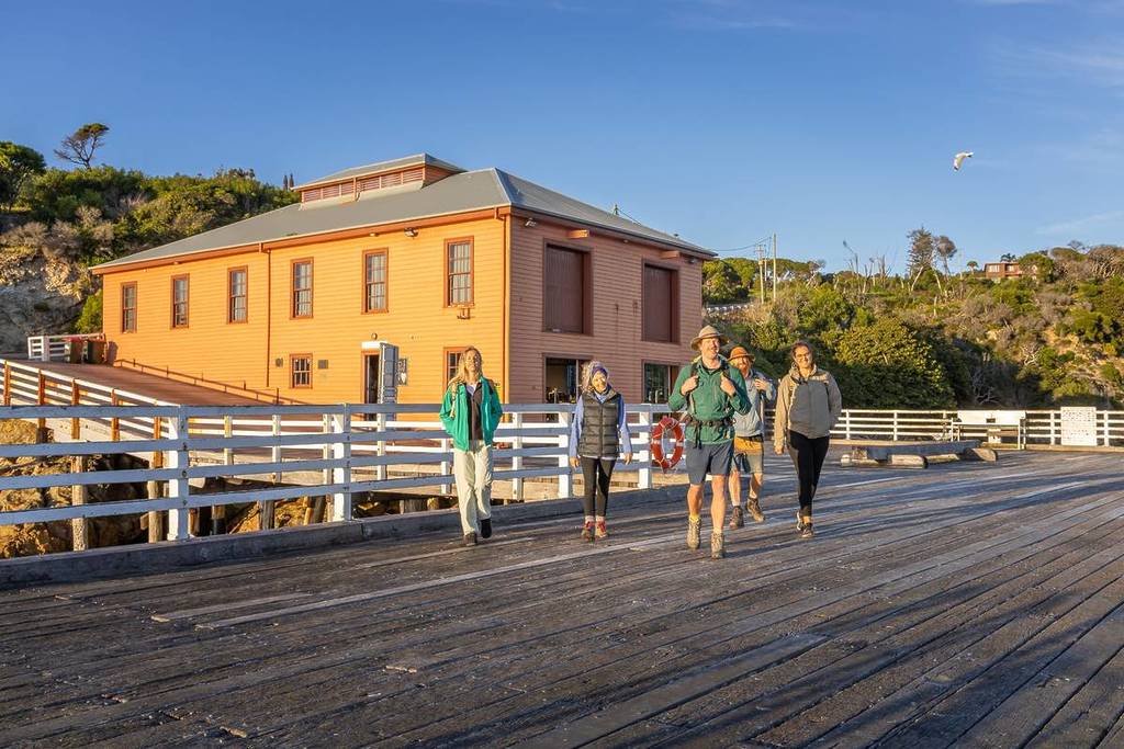 Group of people walking along Merimbula’s historic wharf at sunset, with a large timber building and coastal bushland in the background