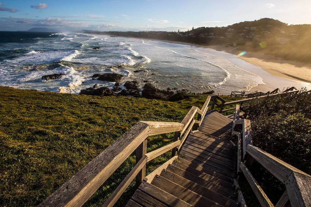 Wooden stairs leading down to a golden beach at Port Macquarie, with waves rolling in and soft afternoon light casting long shadows
