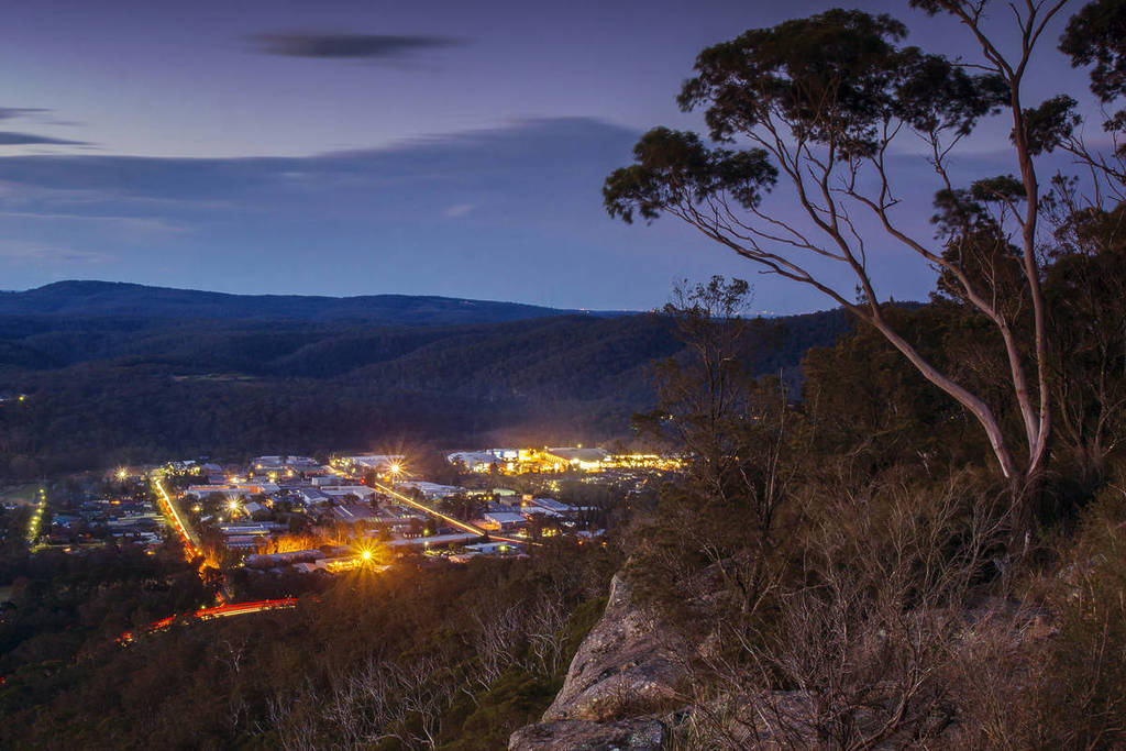 Evening view over Mittagong, NSW, from a rocky bushland lookout, with warm town lights glowing below and silhouetted gum trees framing the scene against a dusky sky
