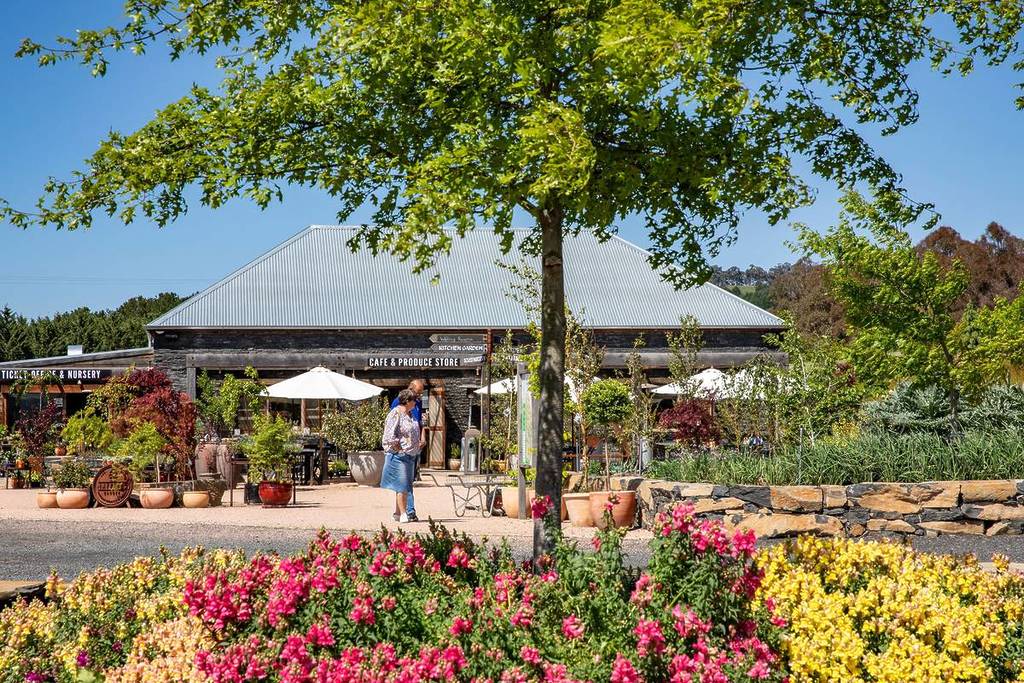 Visitors strolling near vibrant flowers in full bloom in front of the cafe and produce store at Mayfield Garden, set against a clear blue sky and lush greenery on a spring day