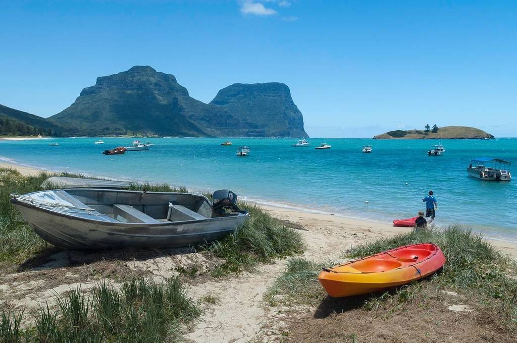 Colorful kayaks and a small boat rest on the sand beside clear turquoise water, with fishing boats anchored offshore and the towering peaks of Lord Howe Island in the distance
