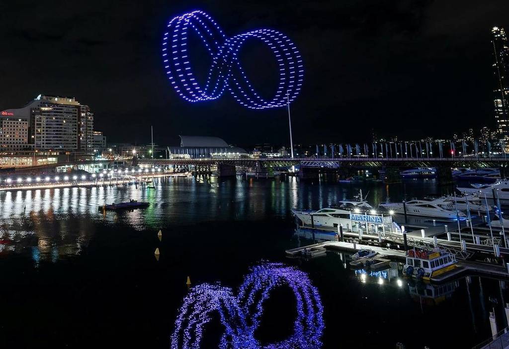 Illuminated drones create glowing blue loops in the night sky above Cockle Bay at Darling Harbour