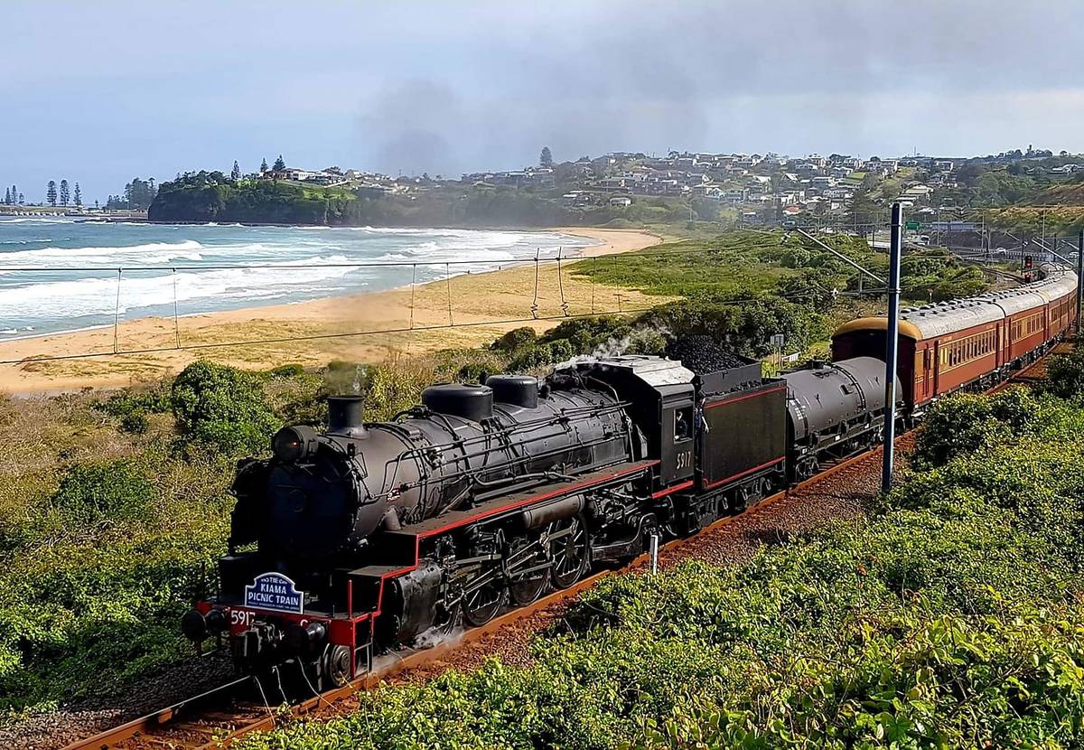 A vintage steam train, numbered 5917, steams along a coastal track near Kiama, passing lush greenery and offering views of the beach and seaside town in the background