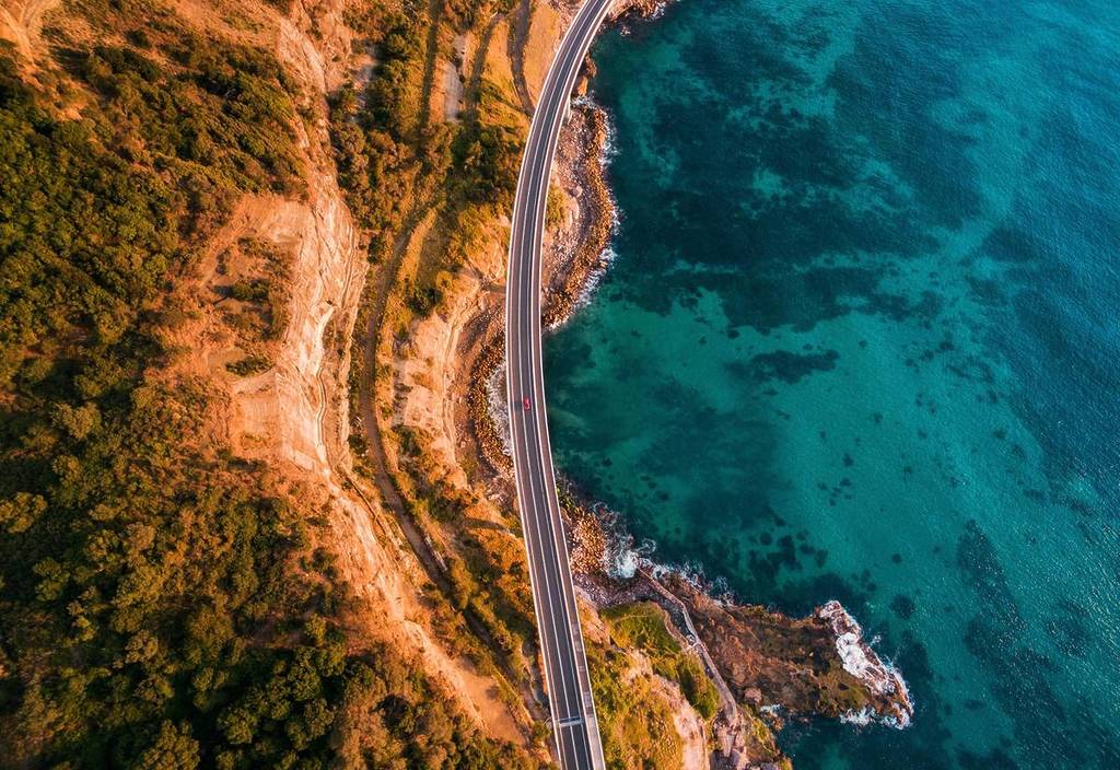 An aerial view of a winding coastal road along the Grand Pacific Drive, with clear turquoise waters on one side and lush greenery on the other