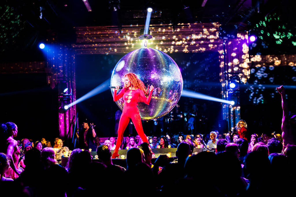 A woman in a red jumpsuit sings in front of a giant disco ball at La Ronde.
