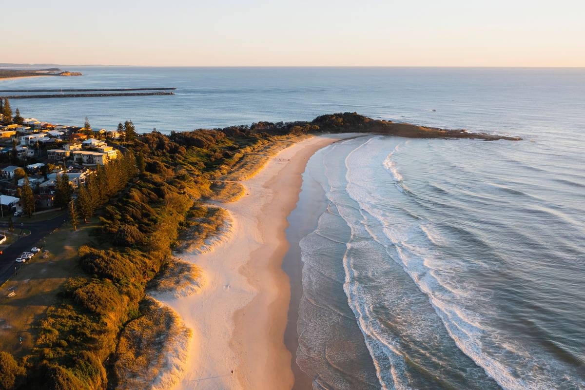 Aerial shot of a beach in Yamba with golden sand, lush greenery, and the town in the background during sunset