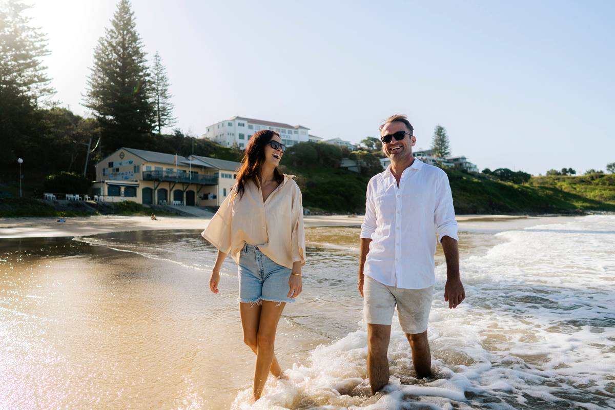 A couple walking along a sandy beach in Yamba, enjoying the waves and sunshine, with buildings in the background