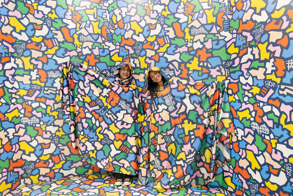 Two friends stand in a camouflage room at Mind Games: Art Alive! wearing giant oodies that have the same pattern as the walls.