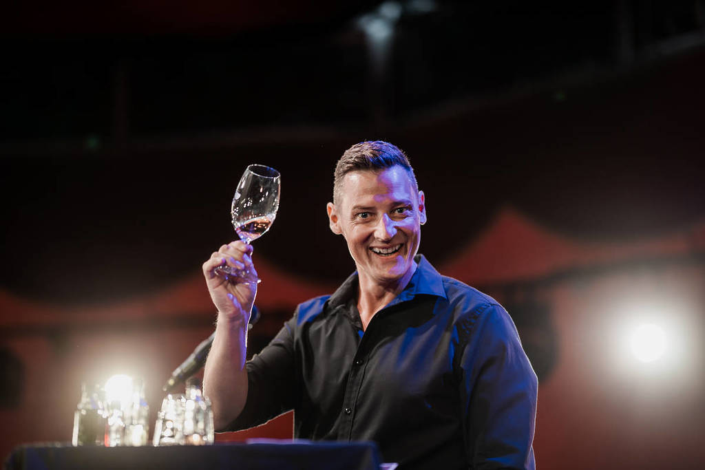 A close-up shot of Merrick Watts holding up a wine glass and smiling at his show, An Idiot's Guide to Wine.