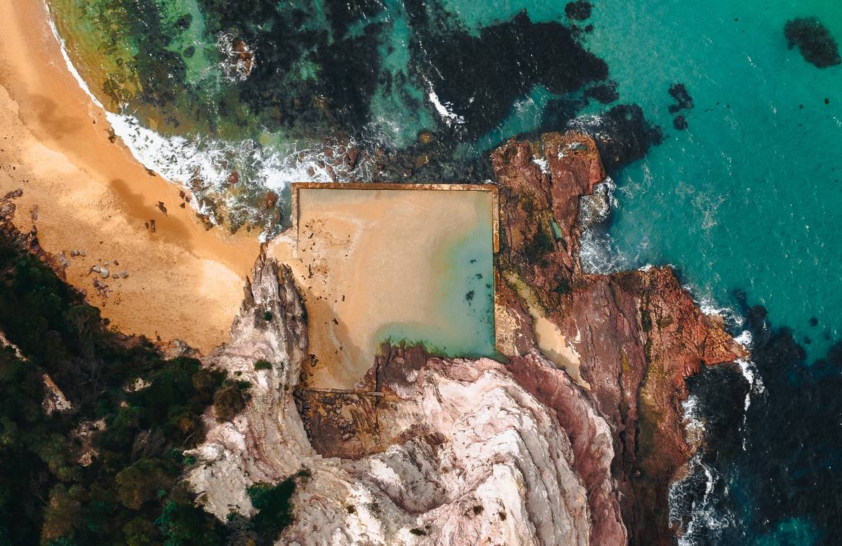 Aerial perspective of Aslings Beach Rock Pool, nestled against pink-hued cliffs and surrounding rocky outcrops