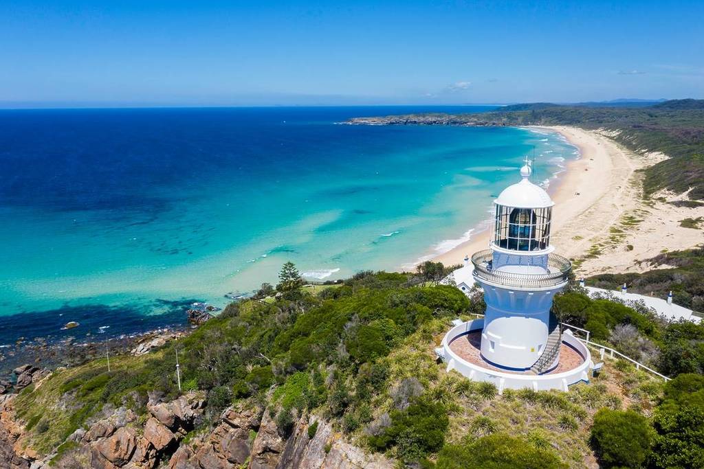 Aerial view of Sugarloaf Point Lighthouse at Seal Rocks, one of NSW's best offbeat coastal towns, perched on a rugged headland overlooking a long stretch of golden sand and bright blue waters