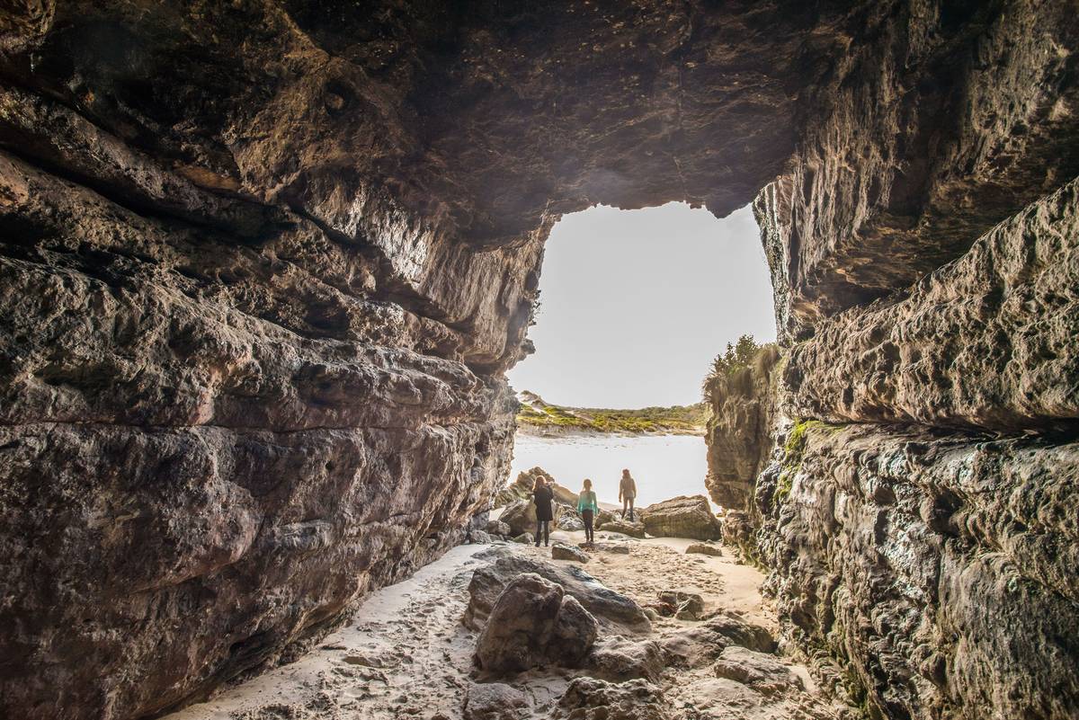 People exploring the natural sea caves at Cave Beach in Booderee National Park, with sunlight streaming through the opening onto sand and rock formations