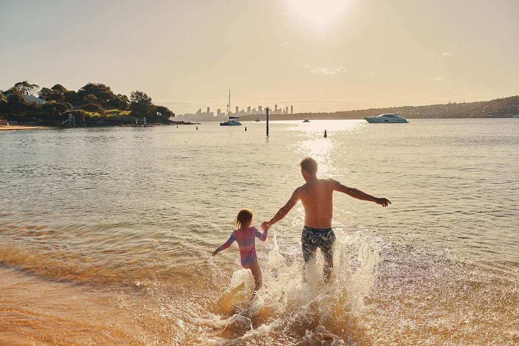 A father and daughter run into the water at a Sydney beach on a golden summer afternoon, with boats anchored and the city skyline in the distance