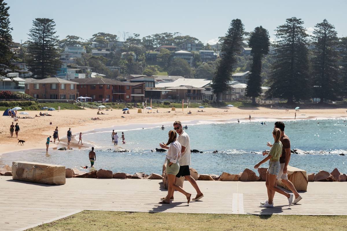 People walking along the scenic Avoca Beach promenade, with the beach and coastal homes in the background