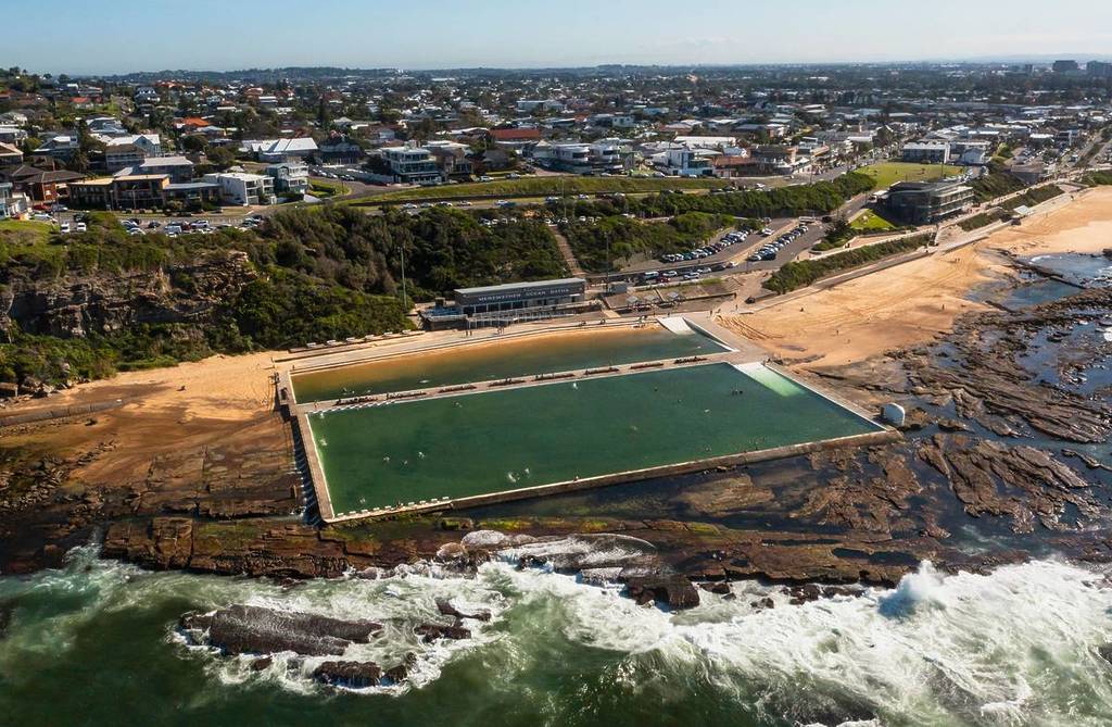 Aerial view of Merewether Ocean Baths in Newcastle, showing the large ocean pool beside the beach, waves breaking over the rocks, and the city visible in the background