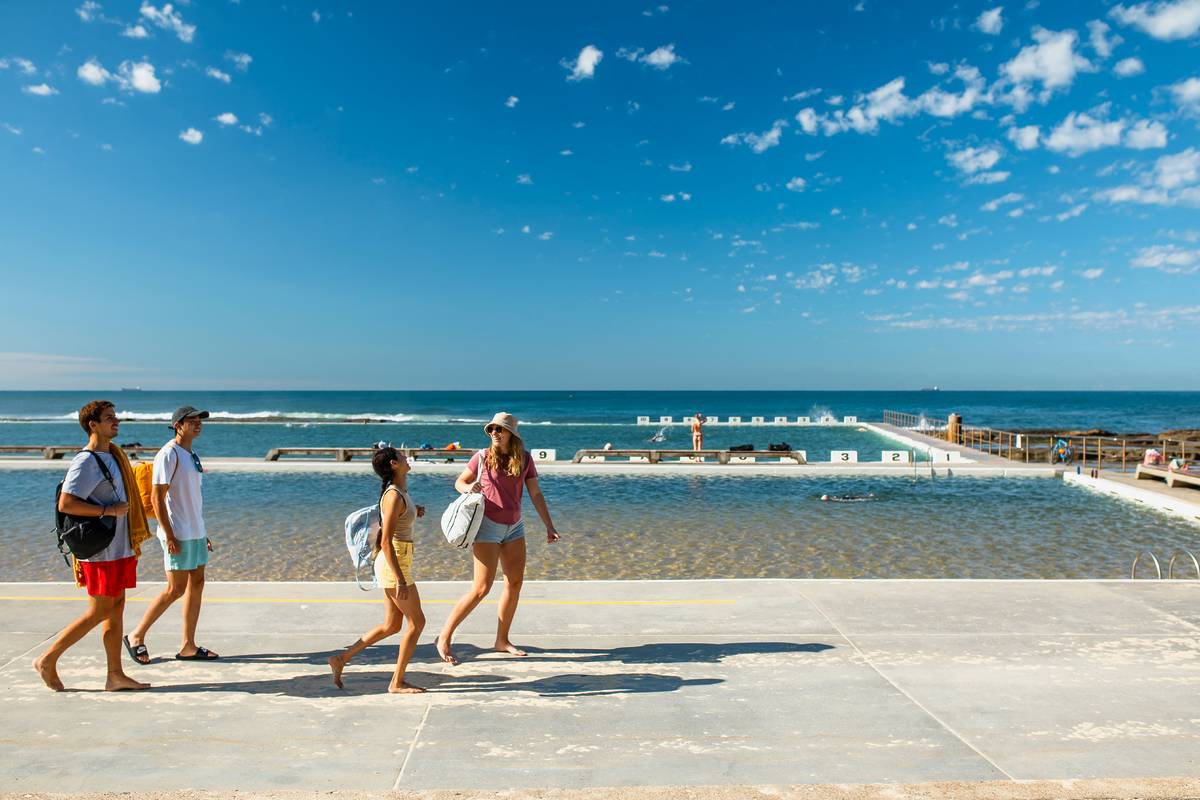 Group of people walking along the edge of Merewether Ocean Baths on a sunny day, with swimmers in the pool and the ocean visible in the distance