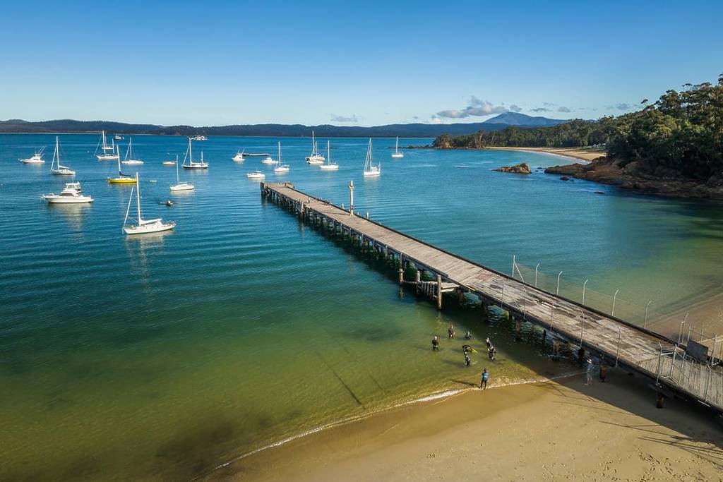 View of Eden Wharf's wooden pier extending over clear blue water, surrounded by anchored boats and gentle coastline