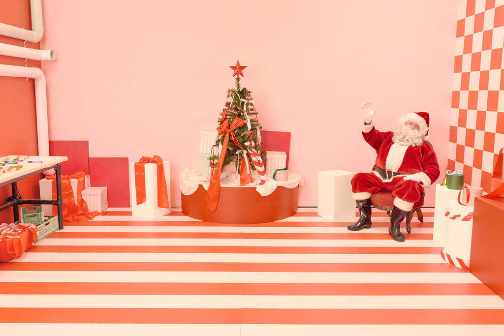 Santa sits on his chair next to the Christmas tree in Santa's Enchanted Photo Workshop and waves.