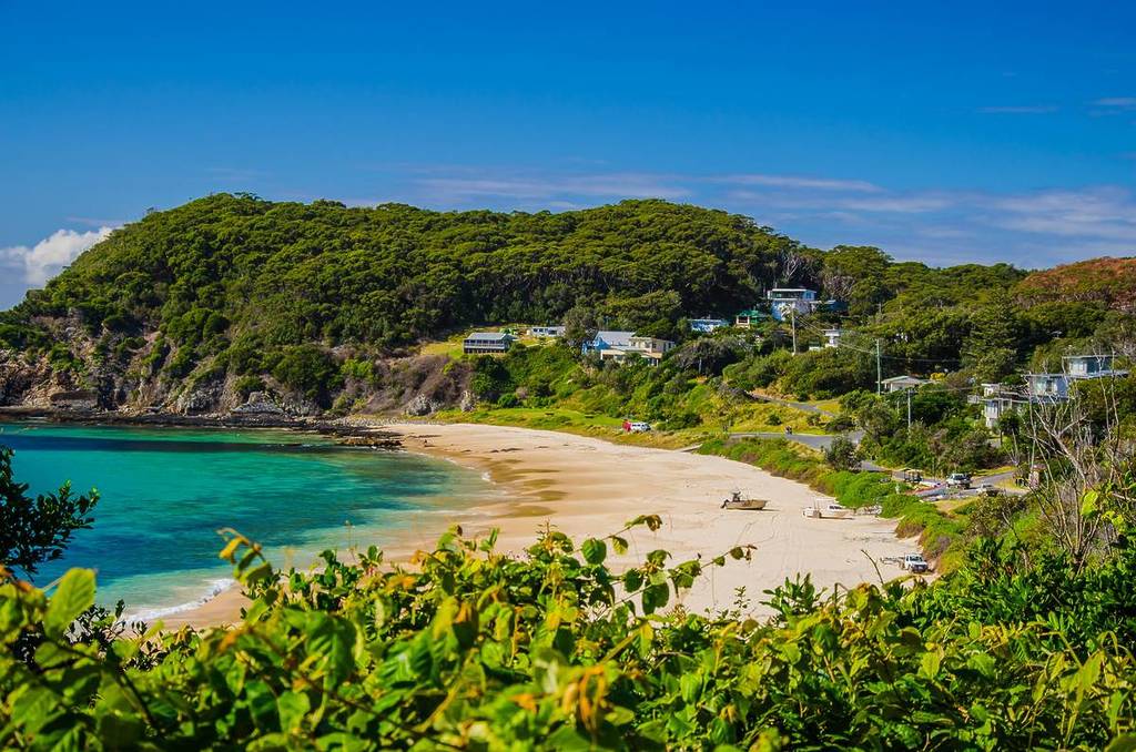 Curved sandy beach with crystal-clear water, backed by a green hillside dotted with coastal homes in Pacific Palms, NSW