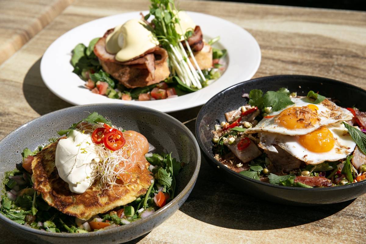 Close-up of three plated breakfast dishes with eggs, greens, and herbs arranged on an outdoor wooden table in Kembali Cafe, NSW