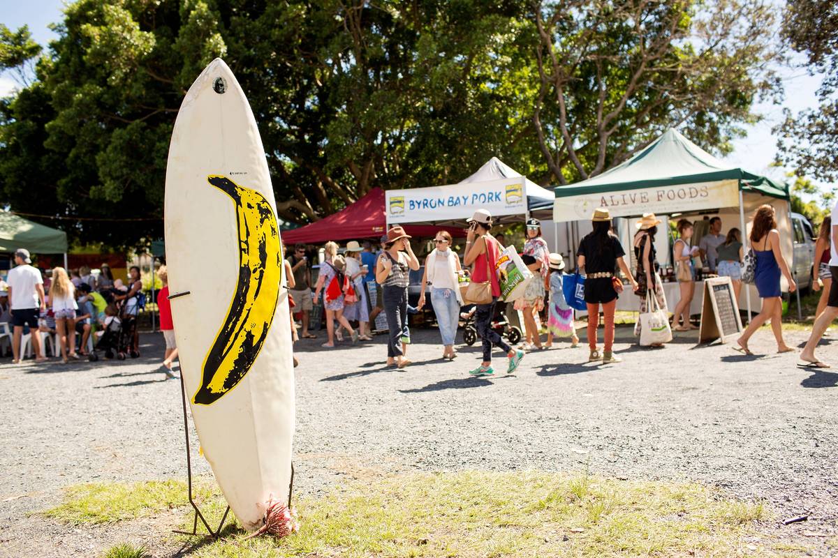 Crowds browsing stalls at the Byron Bay markets with a surfboard display and local food vendors in the background