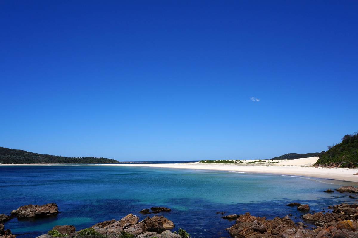 Fingal Bay Beach with a white sand spit connecting to an island, set against blue water and green headlands