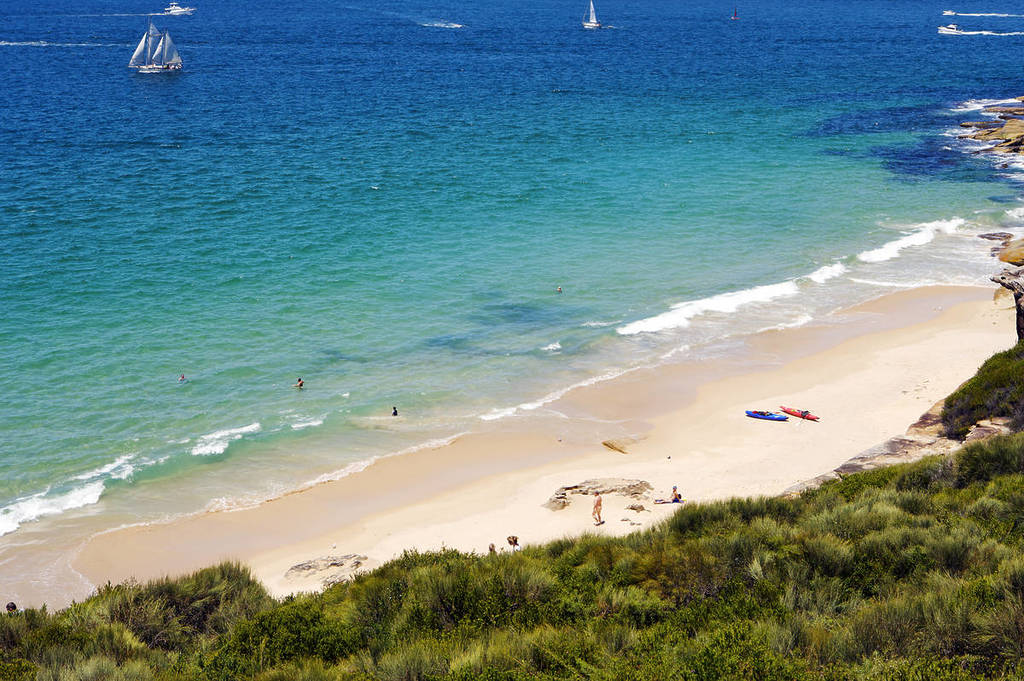 Washaway Beach in Sydney, showing a narrow strip of sand bordered by rock platforms and calm waters, with swimmers and kayaks below