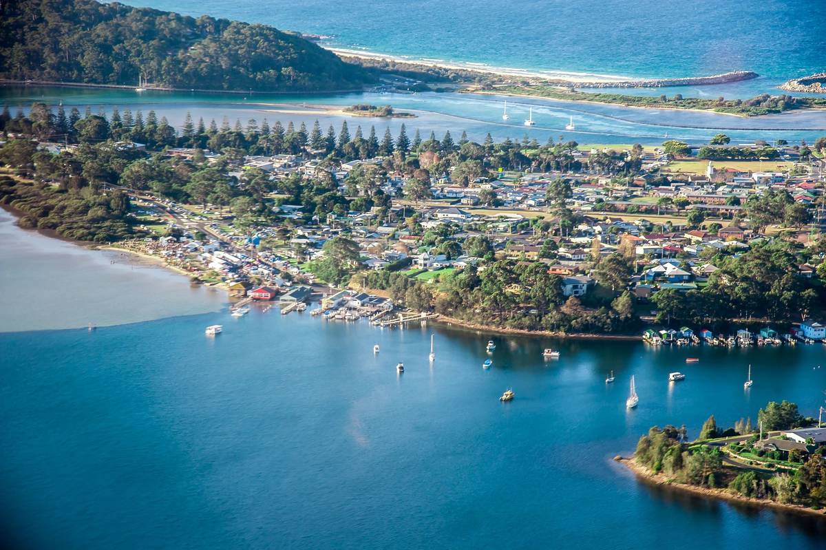 Aerial view of Narooma township overlooking the calm blue waters of Wagonga Inlet, with boats moored along the shoreline and the ocean visible beyond