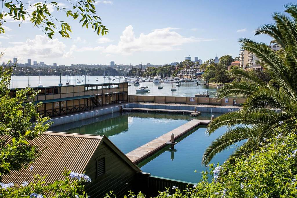 Australia’s oldest swimming pool Dawn Fraser Baths, overlooking Sydney Harbour, with blue water, historic buildings and boats moored nearby