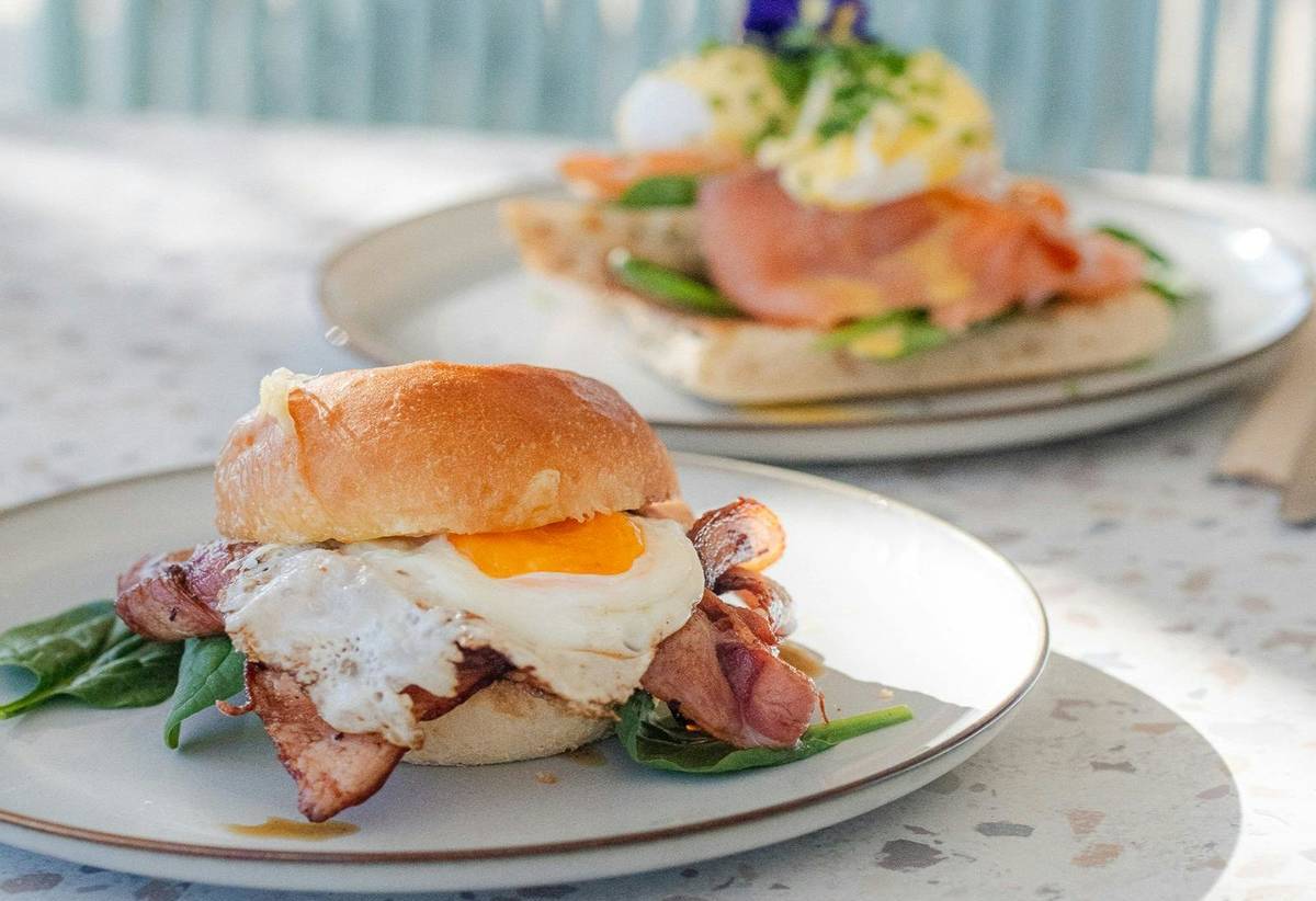 Breakfast burger with bacon, fried egg and spinach on a bun, with avocado toast in the background