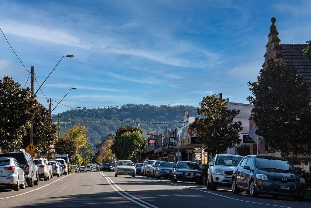 A wide view of Berry's main street lined with shops, parked cars and leafy trees, with green hills rising in the background
