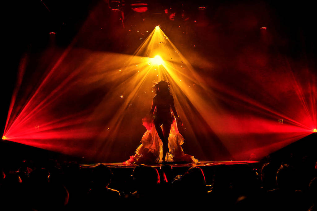 A female performer stands on stage under red and yellow lights in a dark theatre at Late Night Vice. 