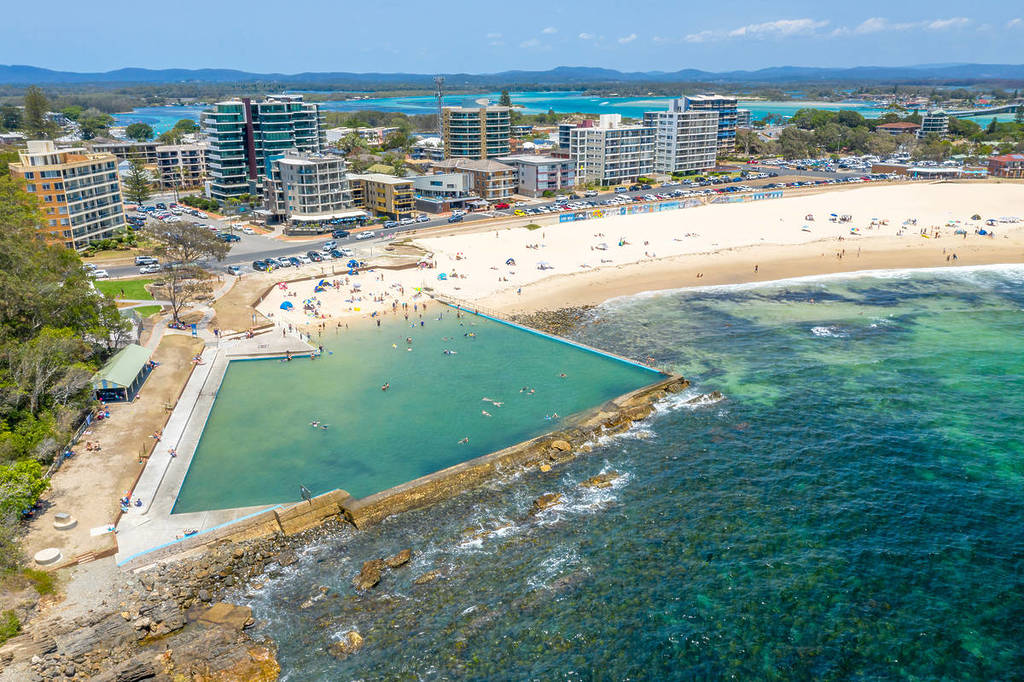 Forster Main Beach ocean baths filled with swimmers, overlooking clear blue waters and sandy shoreline