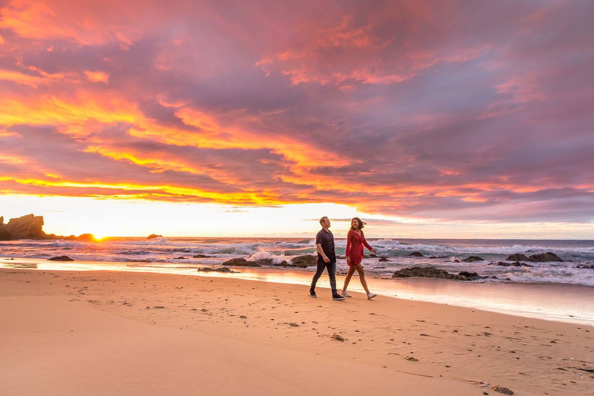 Couple walking along a Bermagui beach at sunset with dramatic orange skies and crashing waves