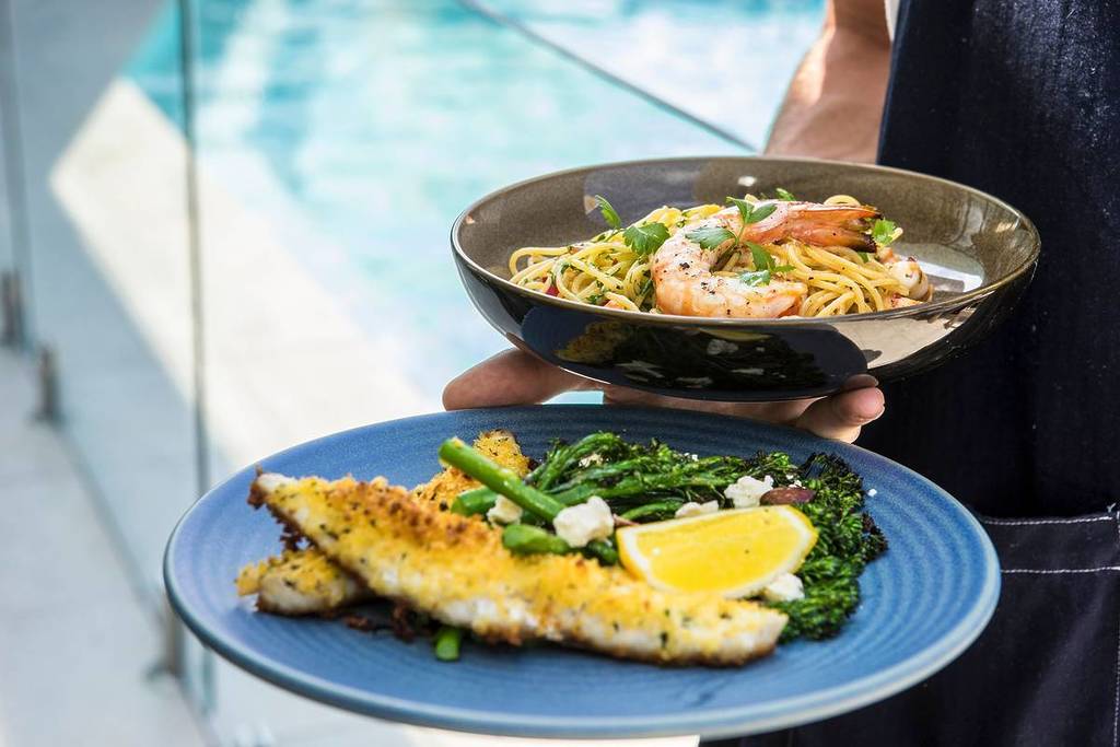 Close-up of seafood dishes being served, featuring grilled fish with greens and lemon alongside prawn pasta, with ocean views in the background in Mollymook